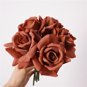 May include: A bouquet of artificial roses in a deep terracotta or burnt orange colour. The flowers feature layered, textured petals with a matte finish, arranged in a dense cluster. The stems are thin, green, and gathered together at the base.
