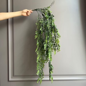 May include: A trailing artificial plant stem featuring numerous small, vibrant green leaves attached to thin, flexible branches. The foliage cascades downwards in a dense, layered arrangement, creating a natural, leafy appearance with varied shades.