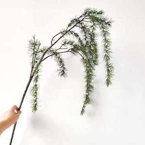 May include: A long, slender artificial branch featuring dark brown bark and multiple cascading stems covered in fine, needle-like green foliage. Small, round, light green textured buds are distributed along the drooping pine-like needles.