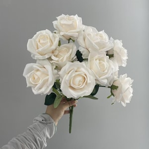 May include: A bouquet of artificial cream-coloured roses featuring multiple full-bloom flower heads with layered petals and dark green leaves attached to a central stem. The roses are arranged in a cluster, showcasing soft off-white tones.