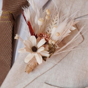 May include: A beige linen jacket with a white and brown dried flower boutonniere. The boutonniere features a large white flower, feathers, and small dried flowers.