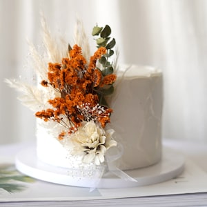 Peut inclure: Un gâteau blanc à la décoration simple est orné de fleurs séchées dans des tons orange et blanc. Le gâteau est sur un support à gâteau blanc et un magazine se trouve en arrière-plan.