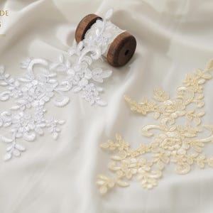 Puede incluir: Dos apliques de encaje floral en blanco y marfil, con una bobina de madera de cinta de encaje blanco. Los apliques presentan intrincados diseños florales y de hojas, perfectos para proyectos nupciales o de artesanía. El fondo es una tela blanca suave.