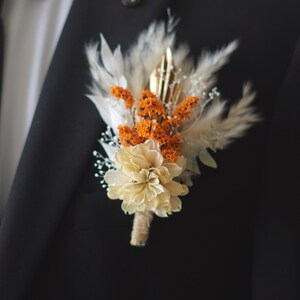 Peut inclure: Une boutonnière de fleurs séchées avec des accents blancs, orange et dorés. La boutonnière est faite de fleurs et d'herbes séchées et est attachée à la boutonnière d'un costume.