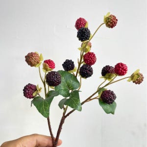 May include: A decorative branch featuring multiple artificial blackberries in various stages of ripeness, ranging from bright red to deep purple and black. The branch includes several green, veined leaves attached to thin, brown, textured stems.