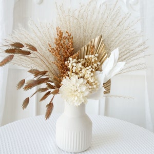 Puede incluir: Un jarrón de cerámica blanco con una superficie texturizada contiene una composición de flores secas. La composición incluye hierba de la pampa blanca, flores blancas secas y hierbas secas marrones.