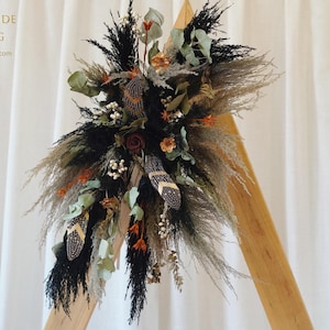 Puede incluir: Arreglo floral decorativo con hierba de la pampa seca, plumas negras y marrones y varias flores secas. El arreglo está colocado contra un marco de madera y un fondo de tela blanca. El texto de la imagen dice "PERFECTBRIDE WEDDING".