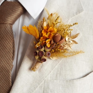 Könnte beinhalten: Nahaufnahme eines beigen Blazers mit einer braunen Strickkrawatte und einer floralen Ansteckblume. Die Ansteckblume zeigt getrocknete Blumen in Gelb-, Orange- und Brauntönen mit goldenen Akzenten. Der Blazer hat eine Tasche.