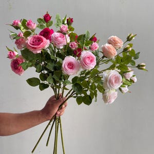 May include: A bouquet of artificial roses in various shades of pink, ranging from soft pastel to deep magenta. The arrangement features multiple blooming flower heads, smaller buds, and green leaves attached to long, thin, green stems.