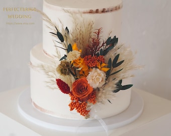 May include: Two-tiered white wedding cake decorated with a floral arrangement. The arrangement includes orange roses, pampas grass, and other dried flowers. The cake is on a white cake stand.