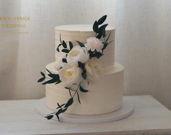 May include: A two-tiered white wedding cake decorated with white flowers and green leaves. The cake is on a white cake stand.