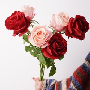 May include: A bouquet of artificial roses featuring three deep red blooms and two soft light pink blooms. The flowers are attached to green stems with green leaves, creating a floral arrangement with contrasting colours and layered petals.