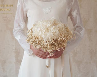 Pode incluir: Um buquê de noiva com flores secas, incluindo hortênsias brancas e pequenas flores de preenchimento bege. O buquê é segurado por uma pessoa vestindo um vestido branco com mangas de renda. O texto "PERFECTBRIDE WEDDING" é visível.