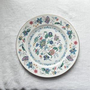 Puede incluir: Plato de cerámica blanco con borde dorado, adornado con un diseño floral y de mariposas en tonos azules, rosas y verdes. El plato presenta un diseño floral central y un borde delicado.