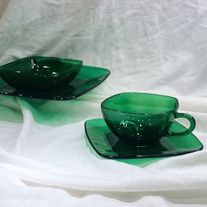 Può includere: Un set di vetreria verde smeraldo, tra cui una tazza da tè con manico e un piattino quadrato, e una ciotola quadrata su un piatto quadrato. La vetreria è realizzata in materiale trasparente e poggia su un tessuto bianco.