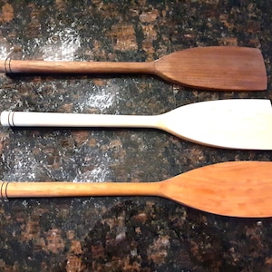 May include: Three wooden spatulas in varying shades of brown and white are displayed. The spatulas have flat, rectangular heads and long, rounded handles. Each handle has a decorative black ring near the end. These kitchen utensils are ideal for cooking and baking.