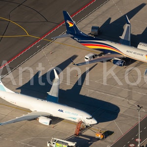 May include: Two airplanes parked on a tarmac. The plane on the left is white with blue accents and says "ASL AIRLINES" on the side. The plane on the right is blue, red, and yellow with "WEST ATLANTIC" on the side.
