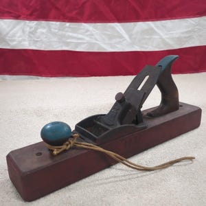 May include: A vintage wooden hand plane with a dark red base and a metal blade assembly. The handle is brown with a teal accent. A blue knob is attached with a tan cord. The year "1921" is etched on the side.