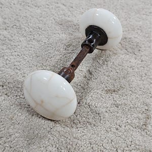 Può includere: Un set di pomelli per porta vintage composto da due pomelli rotondi in ceramica bianca con una sottile texture crepata. I pomelli sono collegati da un perno centrale in metallo marrone scuro e usurato, dotato di una vite a vista per il fissaggio.