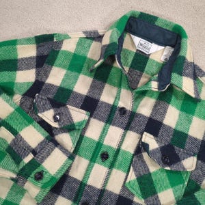 May include: A green, navy, and cream plaid flannel shirt. The shirt has two buttoned chest pockets and a button-down front. The collar is lined with navy fabric. The label reads "Woolrich Woman".