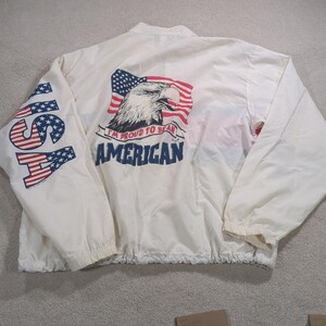 May include: A white windbreaker jacket featuring a graphic on the back of a bald eagle in front of an American flag with the text I'M PROUD TO BE AN AMERICAN. The right sleeve displays the letters USA in a red, white, and blue flag pattern.