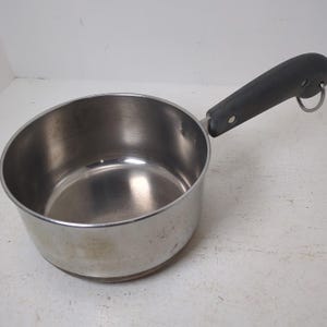 May include: A stainless steel saucepan with a black handle and a metal hanging loop. The pan is round with a slightly flared rim and shows signs of use.