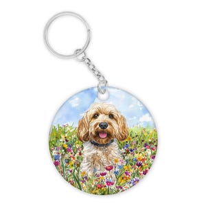 May include: Circular keyring with a watercolour painting of a golden doodle dog in a meadow of vibrant wildflowers. The dog wears a black collar and is smiling. The background shows a blue sky with clouds.