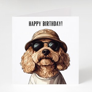 May include: A birthday card with a cartoon dog wearing sunglasses and a tan bucket hat. The dog has curly, light brown fur and is wearing a white t-shirt. The words "HAPPY BIRTHDAY!" are printed in black at the top of the card.