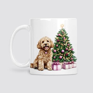 May include: White ceramic mug featuring a light brown dog with a pink collar, sitting next to a decorated Christmas tree with pink baubles and wrapped gifts. The mug has a white handle.