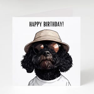 May include: A birthday card with a cartoon illustration of a black dog. The dog wears a beige bucket hat and sunglasses. The text "HAPPY BIRTHDAY!" is printed above the dog's head. The dog is wearing a white t-shirt.
