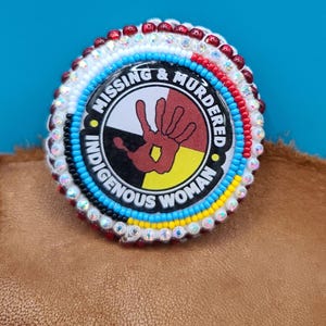 May include: A beaded circle with the words "Missing & Murdered Indigenous Woman" and a handprint in the center. The handprint is red, yellow, black, and white. The circle is surrounded by beads in red, white, blue, and yellow.