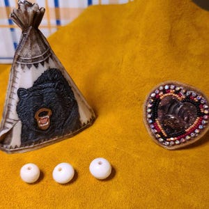 May include: A decorative teepee figurine with a black bear carving, a heart-shaped beaded brooch, and three white beads. The teepee is tan and white with a black bear image. The brooch has a heart-shaped design with red, black, and gold beads.