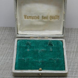 Könnte beinhalten: Eine geöffnete, rechteckige graue Schachtel mit einem grünen Samt-Innenraum. Die Schachtel hat einen Messingverschluss und die Worte "Warranted Best Quality" sind auf der Innenseite des Deckels aufgedruckt.