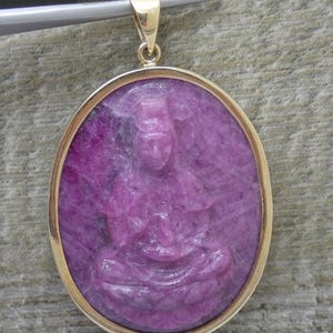 Peut inclure: Un pendentif ovale présentant une figure en pierre précieuse violette et rose sculptée en position assise, sertie dans un cadre en métal doré poli. Le pendentif comprend une bélière dorée au sommet, tenue par des pinces en métal argenté.