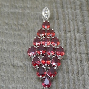 Puede incluir: Colgante de plata con una cascada de piedras preciosas rojas en forma de lágrima. El colgante tiene una pequeña fianza con diamantes en la parte superior. Las piedras están engastadas en metal plateado.