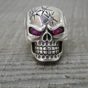 May include: A silver skull ring with two pink gemstone eyes and a starburst design on the forehead.