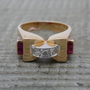 Puede incluir: Un anillo de oro rosa con una barra horizontal central engastada con tres diamantes blancos redondos, flanqueada a cada lado por un rubí rojo de talla cuadrada. La banda tiene un acabado pulido ancho y un diseño geométrico que se estrecha.