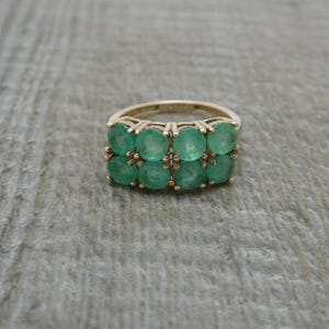 May include: A gold ring featuring two horizontal rows of four round-cut green gemstones each. The eight stones are set in a shared prong setting, creating a rectangular cluster design on the band. The metal has a warm, metallic yellow tone.