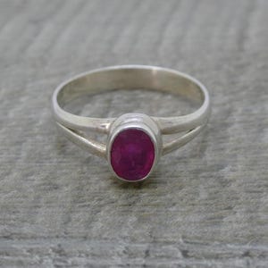 May include: A silver ring featuring a polished, oval-shaped, deep pink gemstone set in a smooth bezel. The band is crafted from a single piece of silver that splits into two separate, slender arms as it approaches the central stone setting.