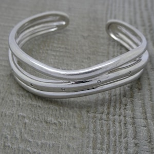 May include: A silver-colored cuff bracelet with a layered design. The bracelet has three parallel bands, with small, evenly spaced embellishments on the middle band. The bracelet is open at the ends and rests on a light gray wooden surface.