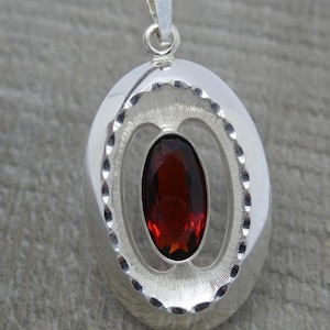 May include: A silver pendant with a red gemstone set in a decorative oval frame.