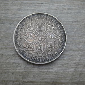May include: A silver Straits Settlements One Dollar coin from 1903. The coin features intricate floral and geometric designs with text in multiple languages. The coin is circular with a detailed edge and is set against a grey wooden surface.