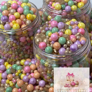 May include: Three glass jars filled with pastel-colored sprinkles, beads, and edible glitter. The jars are on a white surface with a pink and white logo that says "Uptown Sweets, Love baked in every bite!"