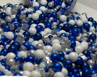 May include: A close-up of a jar filled with blue, white, and silver sprinkles. The sprinkles are a mix of small balls, large balls, and small squares. The jar is tipped over and the sprinkles are spilling out.
