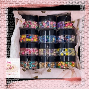 May include: A collection of 12 small jars filled with colorful sprinkles and edible glitter. The jars are arranged in four rows of three. The jars are all black with clear lids. The sprinkles are a variety of colors, including pink, blue, green, purple, gold, and silver.