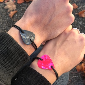 Può includere: Due braccialetti abbinati, ciascuno con un ciondolo a forma di plettro per chitarra. Un plettro è grigio marmorizzato, l'altro rosa acceso. Entrambi sono infilati su cordini neri, indossati ai polsi.