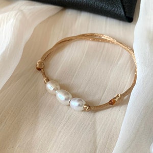 Könnte beinhalten: Ein zartes Armband aus beigefarbenem Band mit drei weißen Perlen.