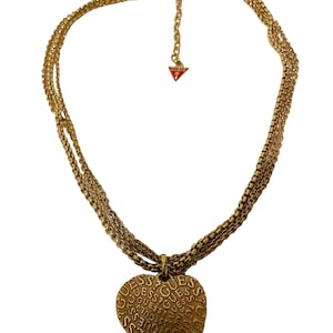 May include: A gold-tone layered chain necklace with a heart-shaped pendant. The pendant is engraved with the word "GUESS" repeatedly.