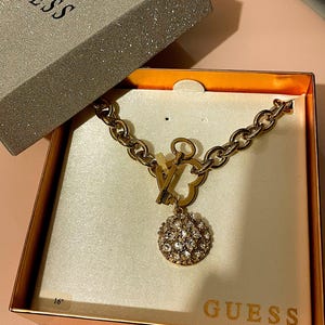 May include: A gold chain necklace with a heart-shaped clasp and a round pendant encrusted with clear crystals. The necklace is in a box with the word "GUESS" printed on it. The box is 40.6 cm long.
