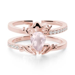 May include: Rose gold ring with a frog motif and a pear-cut, pale pink gemstone. The ring has a double band, with small white gemstones set along the top band. The inscription "G18K" is visible on the lower band.